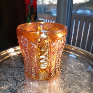 May include: A vintage amber glass pitcher with a diamond-shaped handle and intricate patterns. The pitcher has a iridescent finish and is sitting on a silver tray.
