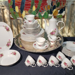 May include: A white porcelain dinnerware set featuring pink rose patterns and silver trim. The collection includes a teapot, creamer, oval platter, bowls, and multiple teacups with saucers, all arranged on a large, round, silver-toned metal serving tray.