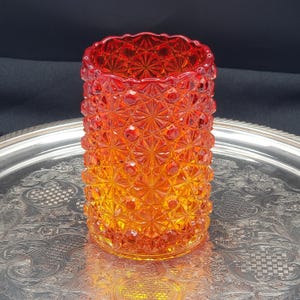 May include: A cylindrical glass vase featuring a vibrant orange and red gradient colour scheme. The exterior surface is covered in a raised, repeating geometric starburst and dot pattern that creates a textured, multifaceted appearance.