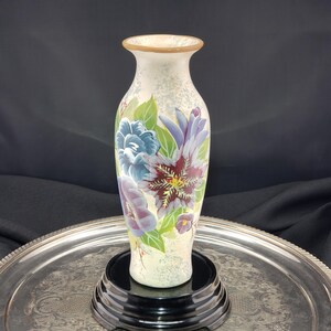 Peut inclure: Un vase en verre haut et élancé avec une base de couleur crème présentant une texture mouchetée. La surface est décorée de motifs floraux peints à la main dans des tons de bordeaux profond, lavande douce et bleu, rehaussés de feuilles vertes.