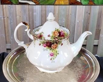 May include: A white porcelain teapot featuring a scalloped edge and a floral pattern of red, yellow, and pink roses with green leaves. The teapot has a matching lid with a pointed finial, a curved handle, and a spout, all accented with gold trim.