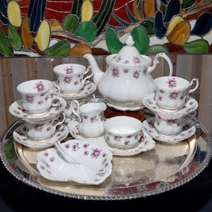Peut inclure: Un service à thé en porcelaine blanche orné d'un délicat motif floral violet avec des bordures dorées. L'ensemble comprend une théière avec couvercle, quatre tasses avec soucoupes, un pot à lait, un sucrier et un petit plat, disposés sur un plateau.