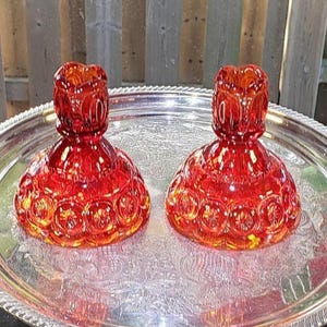 Peut inclure: Deux bougeoirs en verre rouge rubis assortis présentant un motif pressé de lune et d'étoiles. Chaque pièce possède une base large et arrondie qui se rétrécit vers le haut jusqu'à un bord festonné. Le verre est translucide et éclatant.