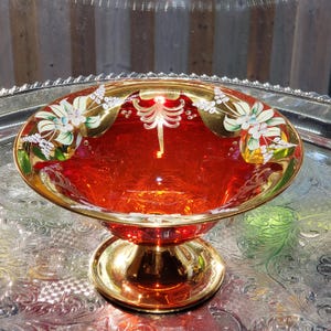May include: A deep red glass pedestal bowl featuring a wide gold rim and base. The bowl is decorated with hand-painted white floral motifs with green leaves and gold filigree accents. The glass has a smooth, polished finish and a flared shape.