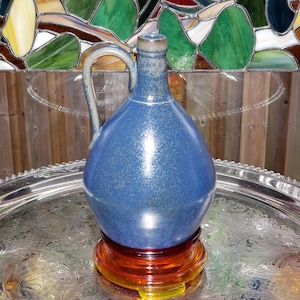 May include: A blue ceramic jug with a handle, resting on a decorative amber-colored stand. The jug has a rounded body and a narrow neck. The surface of the jug has a speckled appearance. The jug is displayed on a silver tray.