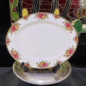 May include: An oval white porcelain serving platter featuring a scalloped edge with gold trim and a floral pattern of red and yellow roses. The platter is displayed on a round silver-toned metal tray with an intricate embossed pattern.