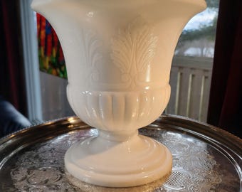 May include: A white milk glass vase featuring a wide flared rim, a rounded body with vertical fluted detailing, and an embossed leaf pattern around the upper section. The vase stands on a circular pedestal base with a smooth, glossy finish.