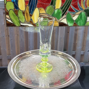 May include: A clear glass with a green base and a yellow-green stem sits on a silver tray with an ornate design.
