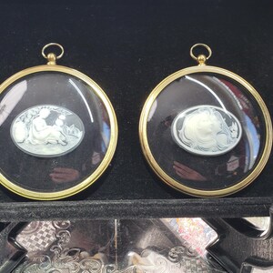 May include: Two circular wall hangings featuring gold-toned metal frames and glass covers. Each contains an oval cameo with a white relief figure set against a black background. The left cameo depicts a seated figure, while the right shows a profile.