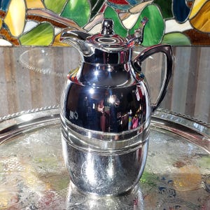 May include: A polished silver carafe with a curved handle and a lid, reflecting the surroundings. It sits on a silver tray with a decorative edge. The background features a stained glass window.