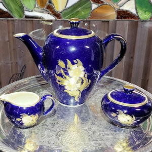 May include: A cobalt blue porcelain tea set comprising a teapot, a creamer, and a lidded sugar bowl, all adorned with gold floral patterns and gold rim accents, displayed on a round, ornate silver-toned metal serving tray.
