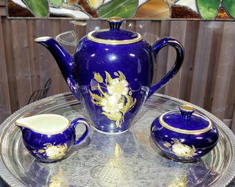 May include: A cobalt blue porcelain tea set featuring a teapot, a creamer, and a lidded sugar bowl, all decorated with gold floral patterns and gold rim accents, arranged on a round, ornate silver-toned metal serving tray.