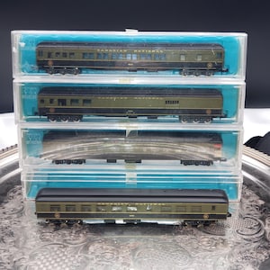 May include: Four model train passenger carriages in olive green and black livery with Canadian National lettering. Three carriages are enclosed in individual clear plastic display cases with blue inserts, while one carriage sits outside in front.