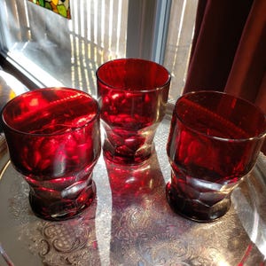 Peut inclure: Trois verres à boire assortis en verre rouge rubis avec une base géométrique à facettes. Chaque verre présente un bord lisse et arrondi ainsi qu'une partie inférieure texturée et épaisse qui capte la lumière, créant des reflets cramoisis.