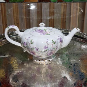 May include: White porcelain teapot with a lid, handle, and spout. The teapot is decorated with purple floral patterns and green leaves. The teapot is sitting on a silver tray.
