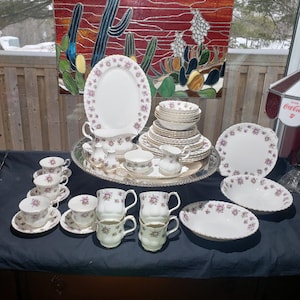 Peut inclure: Service à thé et dîner en porcelaine vintage. L'ensemble comprend tasses, soucoupes, mugs, bols, assiettes et plats de service. Chaque pièce blanche est ornée d'un motif floral délicat violet et vert, avec des bordures dorées.