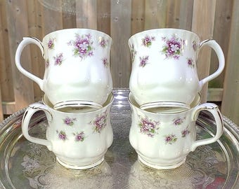 May include: A set of four white ceramic mugs featuring a repeating pattern of small purple and green floral clusters. Each mug has a scalloped base, a curved handle, and a gold-toned rim. The mugs are arranged in two pairs, one stacked on the other.