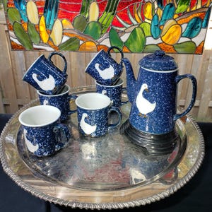 May include: A vintage blue speckled enamelware coffee set with a white goose design. The set includes a coffee pot and six mugs, all displayed on a silver serving tray. The background features a colorful stained-glass window.