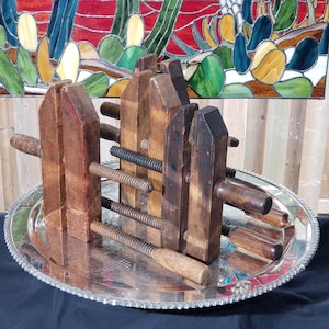 May include: A collection of antique wooden clamps with threaded rods and handles, arranged on a silver-colored, oval tray. The clamps are various sizes and show signs of age and use. A stained glass window is in the background.