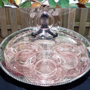 May include: A set of pink glass plates featuring etched floral patterns and a central starburst design. The collection includes several circular plates arranged on a large, round, silver-toned metal tray with a decorative rope-style edge.