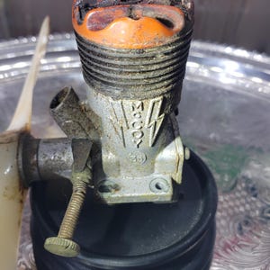 May include: A vintage McCoy 35 glow engine for model aircraft, featuring a metallic silver body with a prominent orange cylinder head. The engine casing displays the embossed McCoy brand name flanked by lightning bolt graphics and the number 35.