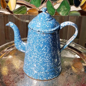 May include: A blue and white speckled enamel coffee pot with a matching lid, a curved spout, and a side handle. The metal pot features a mottled pattern throughout its exterior surface, including the lid, handle, and spout, with a glossy finish.
