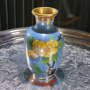 May include: A small cloisonné vase featuring a light blue background with a repeating cloud pattern. The vase is decorated with a large yellow flower, green leaves, and a dark rock base. It has a gold-toned metal rim at the top and bottom.