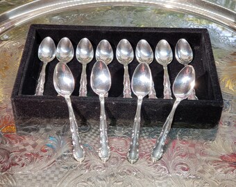 May include: A set of twelve silver-toned metal spoons arranged in a black velvet-lined display box. The spoons feature a polished finish with decorative embossed detailing on the handles. The box is positioned on a large, round, ornate silver tray.
