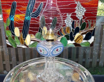 May include: A clear glass oil lamp with a brass burner and a painted blue oval band featuring small red and green floral motifs. Behind the lamp is a rectangular stained glass panel depicting a desert landscape with cacti against a red sky.