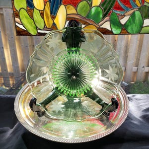 May include: A translucent green glass bowl with a scalloped edge and etched floral patterns, featuring a central sunburst design. The bowl is supported by a black stand and rests inside a round, polished silver-toned metal tray with a decorative rim.