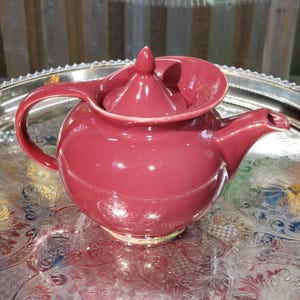 Peut inclure: Une théière en céramique rouge bordeaux brillant avec un corps arrondi, une anse incurvée, un bec court et un couvercle assorti doté d'un bouton pointu. La théière présente une surface lisse et réfléchissante ainsi qu'une large ouverture.