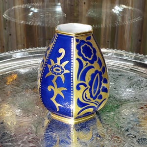 May include: A hexagonal ceramic vase featuring a deep cobalt blue glaze with intricate gold floral patterns and vertical dotted lines. The vase has a white interior rim and a gold-trimmed edge, showcasing a tapered shape with flat panels.