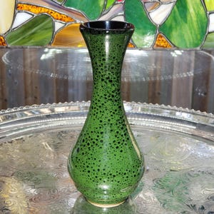 May include: A ceramic vase with a narrow neck and a bulbous base, featuring a vibrant green glaze speckled with numerous small black dots. The interior rim of the vase is finished in a solid dark black glaze, creating a distinct contrast.
