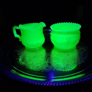 May include: A creamer and a sugar bowl made of glowing neon green uranium glass. Both pieces feature a scalloped rim and a ribbed body design. The creamer has a small handle and a pouring spout. They are positioned on a reflective silver tray.
