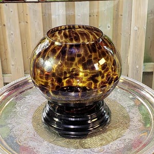 May include: A spherical glass bowl featuring a brown and amber tortoiseshell pattern with a glossy finish. The bowl is supported by a solid black circular pedestal base, creating a distinct contrast between the patterned glass and the dark stand.