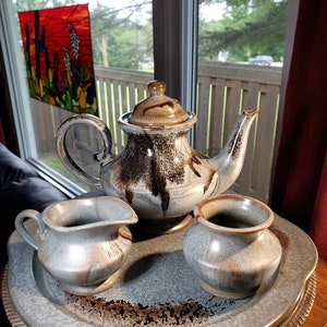 May include: A brown and grey ceramic teapot with a matching creamer and sugar bowl. The teapot has a brown glaze with a speckled pattern. The creamer and sugar bowl have a similar glaze but are a lighter shade of grey. The set is displayed on a silver tray.
