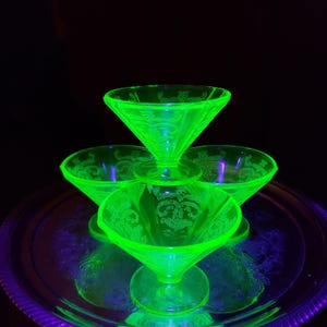 May include: A set of four glowing neon green glass dessert bowls arranged on a silver tray. Each bowl features a wide, flared rim and a short pedestal base, decorated with an intricate, etched floral pattern that fluoresces under UV light.