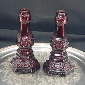 May include: A pair of deep red glass candle holders featuring a pressed pattern with circular and geometric motifs. Each holder has a hexagonal base and a tapered neck, showcasing a translucent finish that catches the light across the surface.
