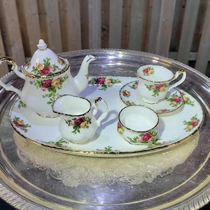 Peut inclure: Un service à thé en porcelaine blanche orné d'un motif floral de roses rouges et jaunes avec des bordures dorées, comprenant une théière, un pot à lait, un sucrier, une tasse et sa soucoupe, disposés sur un grand plateau ovale argenté.