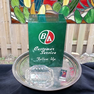 May include: A green metal box with a white "BA" logo and the text "Customer Service Follow Up". The box is sitting on a silver tray with a glass ashtray and a silver lighter.