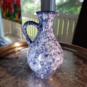 May include: A ceramic pitcher featuring a bulbous body and a single handle, decorated with a mottled blue and white sponge-painted pattern. The vessel has a narrow neck that flares slightly at the rim, creating a distinct pouring spout.
