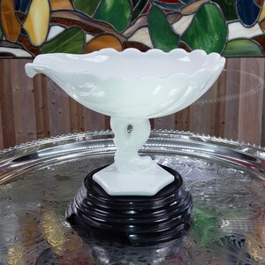May include: A white milk glass compote dish featuring a scalloped rim and a pedestal base shaped like a dolphin or fish. The bowl has a smooth, opaque finish and rests on a black circular stand, showcasing a detailed sculptural design.
