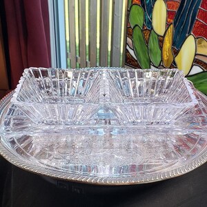 May include: A clear glass divided serving dish with two square compartments featuring a faceted, geometric pattern. The dish rests on a round, silver-toned metal serving tray with an ornate, embossed floral design and a decorative rope-style edge.
