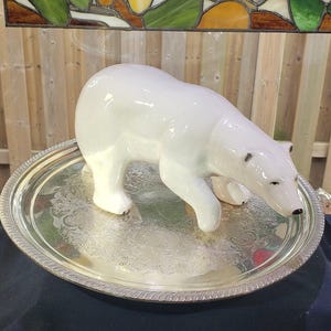 May include: A white ceramic polar bear figurine standing on a round, silver-toned metal tray. The tray features an intricate, embossed floral pattern along its center and a decorative, ridged edge. The polar bear has a smooth, glossy finish.