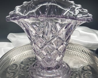 May include: A light purple glass vase featuring a diamond-cut pattern on the body and a scalloped, ruffled rim. The vase has a wide, flared opening that tapers down to a circular base, showcasing a translucent finish and intricate pressed glass details.