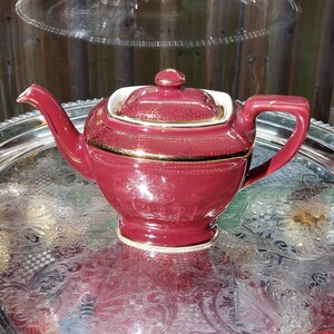 May include: A deep red ceramic teapot featuring a gold crackle pattern on the lid and upper body. The teapot has a curved spout, a sturdy handle, and a matching lid with a small knob. The finish is glossy with gold trim accents.