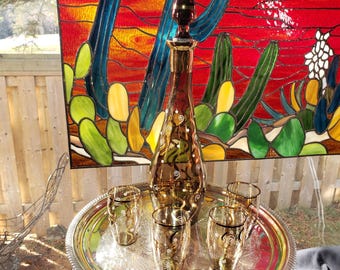 May include: A tall amber glass decanter with a matching stopper and three small glasses, all featuring white swirl patterns. The set sits on a round, polished silver-tone metal tray with an ornate embossed border and center design.