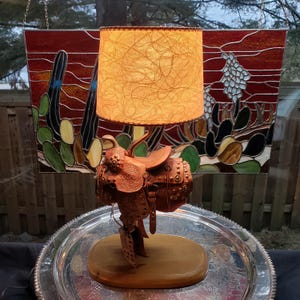 May include: A table lamp featuring a miniature brown leather saddle as the base, set on a smooth oval wooden platform. The lamp has a cylindrical shade with a textured, amber-coloured fibrous material that emits a warm, soft glow when illuminated.