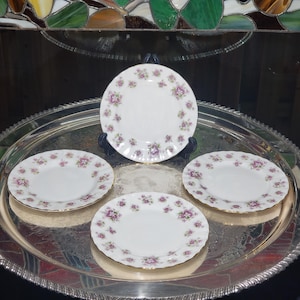May include: A set of four white porcelain plates featuring a delicate floral pattern with small pink and purple flowers and green leaves arranged around the rims. The plates have scalloped edges with gold trim and are displayed on a silver tray.