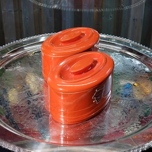 May include: Two orange ceramic containers with oval lids sit on a silver platter. The containers have a glossy finish and an oval handle on the lid. The platter features an ornate, engraved design. The containers are likely for storage.